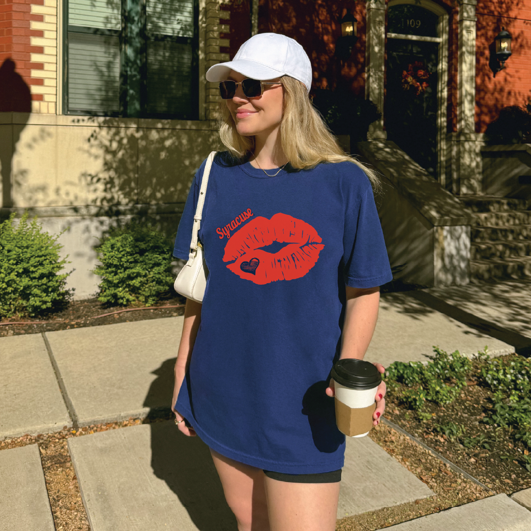 Woman wearing navy t-shirt with bold red kiss graphic and Syracuse script, styled casually with a coffee cup, crossbody bag, and sunglasses for game day streetwear