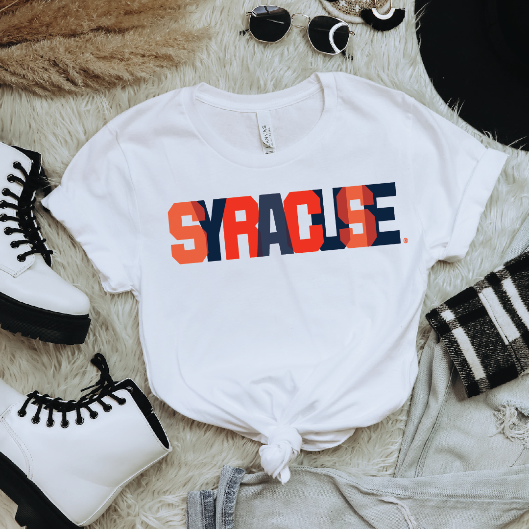 Flatlay of white t-shirt with bold orange and navy block "SYRACUSE" graphic, styled with white combat boots, denim shorts, and sunglasses for a cool streetwear look