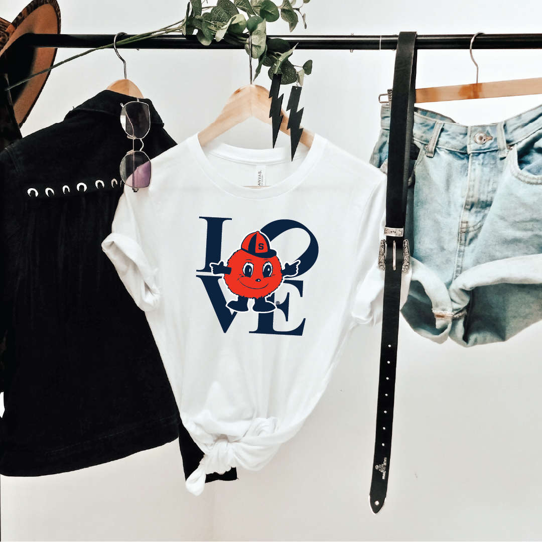 White t-shirt featuring bold navy stacked "LOVE" design with Otto the Orange mascot, styled on hanger with denim shorts, belt, and jacket for casual game day look