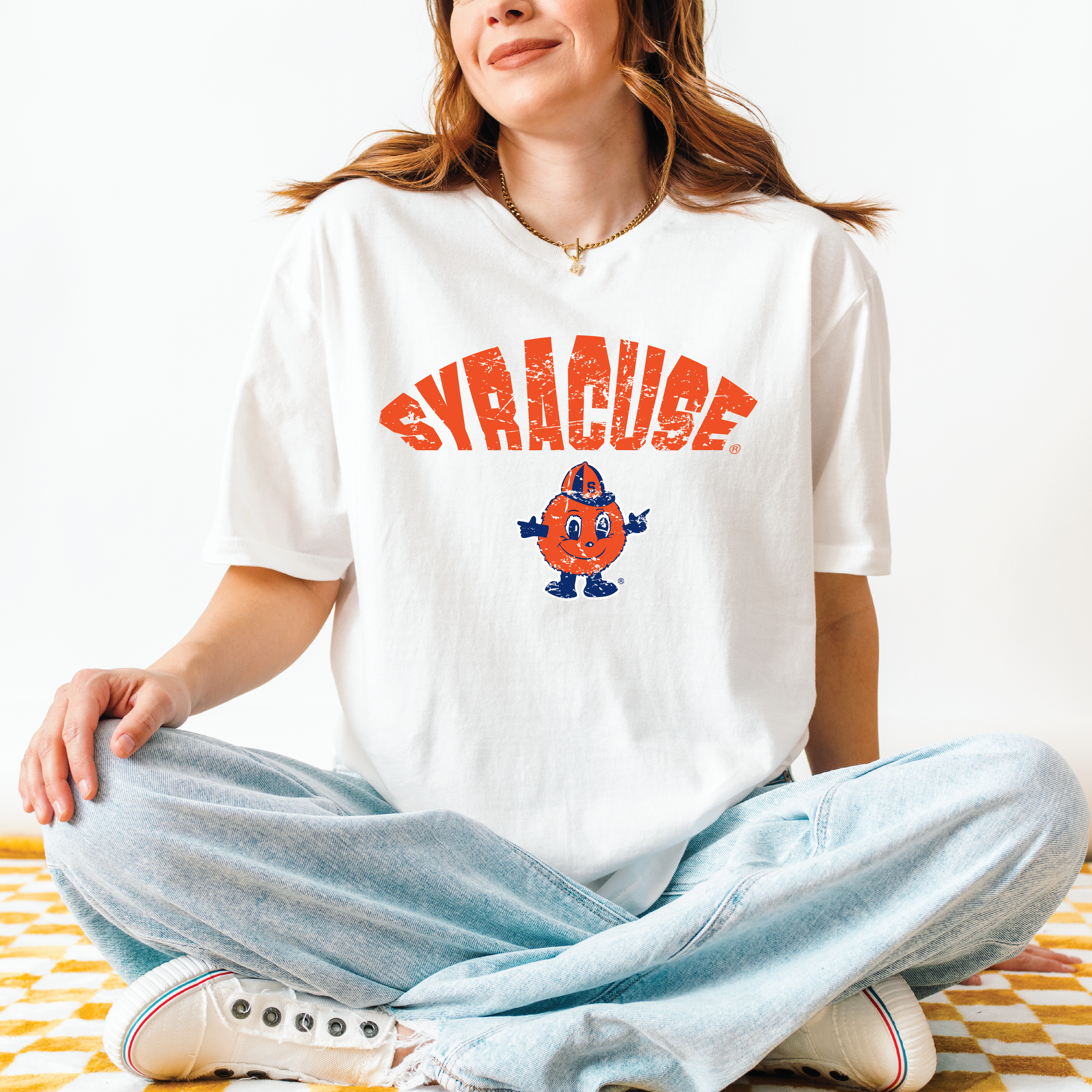Model wearing white t-shirt with distressed red “SYRACUSE” arch and full-color Otto the Orange mascot – sitting cross-legged on yellow checkered blanket