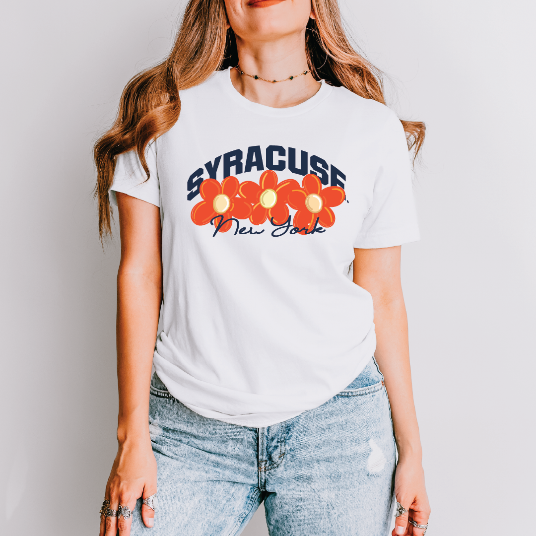 Woman wearing white t-shirt with bold navy “Syracuse” arch, bright orange flowers, and “New York” script – styled casually for a retro game day look